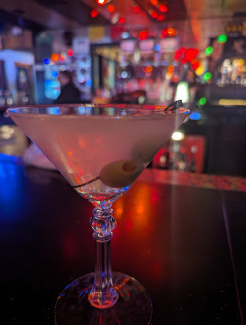 A close-up of a martini glass with an olive, set against a colorful, blurred bar background.