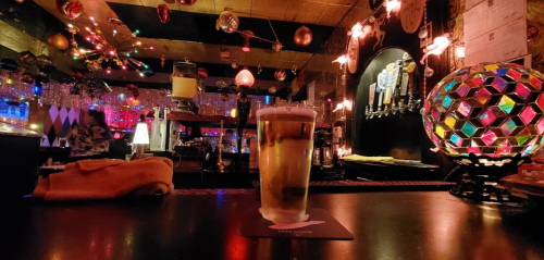 A glass of beer sits on a bar counter, surrounded by colorful lights and festive decorations in a lively pub.