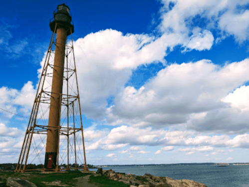 A tall lighthouse stands on rocky shore under a blue sky with fluffy white clouds, overlooking the water.