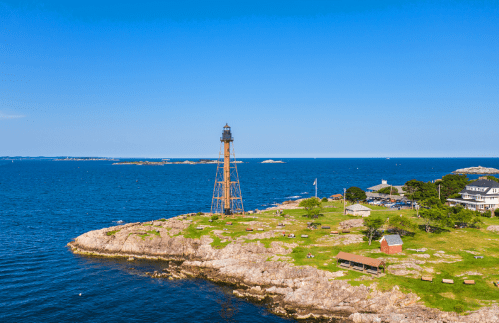 A scenic view of a lighthouse on a rocky shore, surrounded by blue water and green grass under a clear sky.