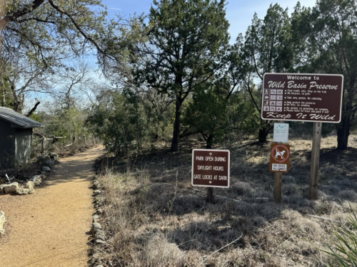 Pathway leading to Wild Basin Preserve, with signs about park hours and rules, surrounded by trees and natural scenery.