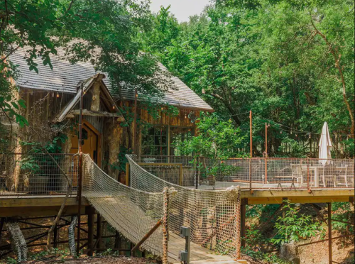 A rustic cabin surrounded by trees, featuring a wooden deck with a net bridge and outdoor seating.