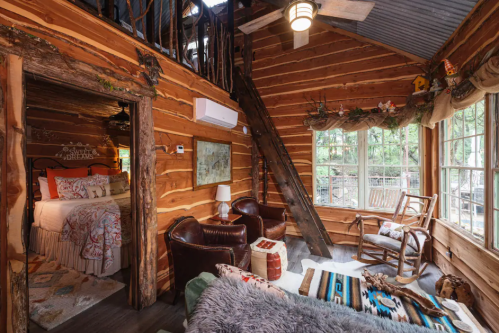 Cozy log cabin interior with wooden walls, a loft, and rustic furniture, featuring a bed, chairs, and natural light.