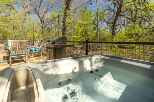 A hot tub on a patio surrounded by trees, with a grill and outdoor seating in the background.