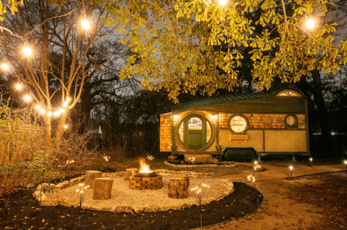 A cozy cabin surrounded by trees, string lights, and a fire pit, creating a warm, inviting atmosphere at night.