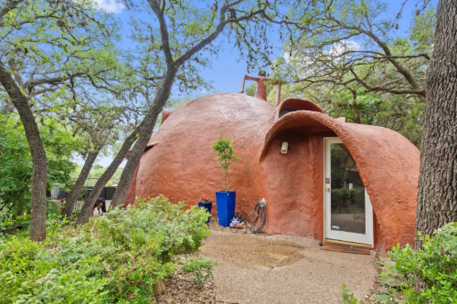 A unique, rounded adobe-style home surrounded by trees and greenery, featuring a curved entrance and a blue container.