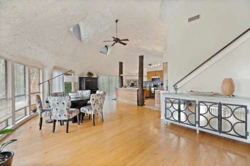 Spacious living area with a dining table, modern kitchen, and large windows, featuring unique ceiling design and wooden accents.