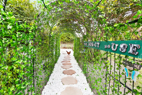 A garden path with stone steps leads through a green archway covered in vines, with a sign reading "Hobbit House."