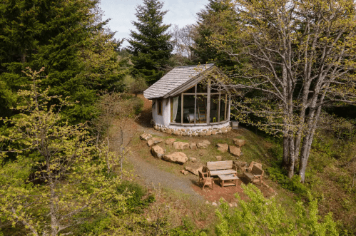 A cozy cabin with large windows, surrounded by trees and rocks, featuring a small outdoor seating area.