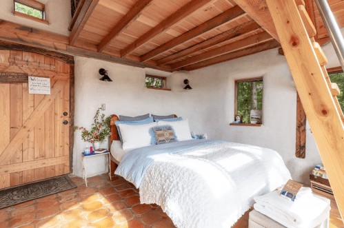 Cozy bedroom with a wooden bed, natural light, and rustic decor, featuring a welcoming door and potted plants.