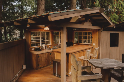 Cozy outdoor kitchen with wooden counters, a sink, and seating, surrounded by trees. Warm lighting adds a welcoming touch.