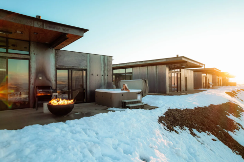 Modern cabins with large windows, a hot tub, and a fire pit, surrounded by snow under a clear sky at sunset.