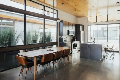 Modern kitchen and dining area with large windows, wooden ceiling, sleek appliances, and a minimalist table setting.