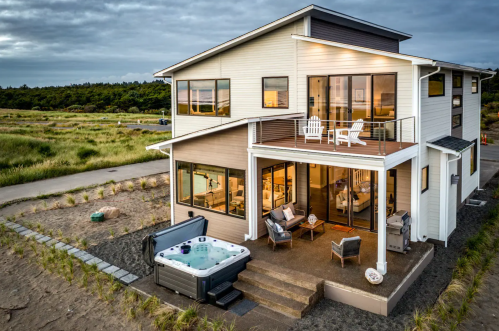 Modern two-story house with large windows, a deck, and a hot tub, surrounded by grassy landscape under a cloudy sky.