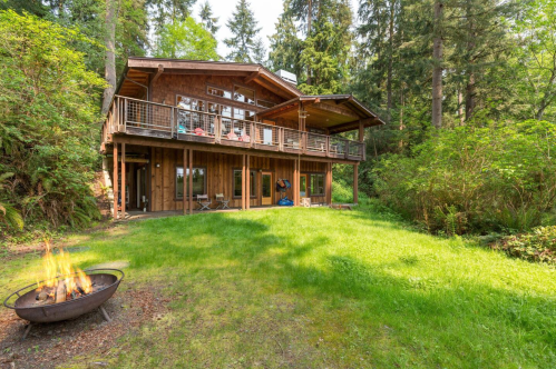 A wooden cabin surrounded by trees, featuring a fire pit in the foreground and a spacious deck overlooking the greenery.