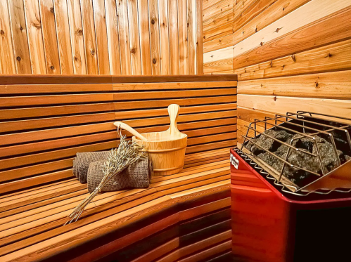 A wooden sauna interior with benches, a wooden bucket, towels, and a stove with rocks.