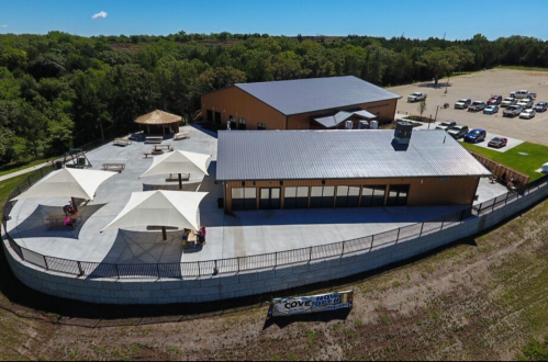 Aerial view of a large building with a patio, shaded areas, and a parking lot surrounded by greenery.