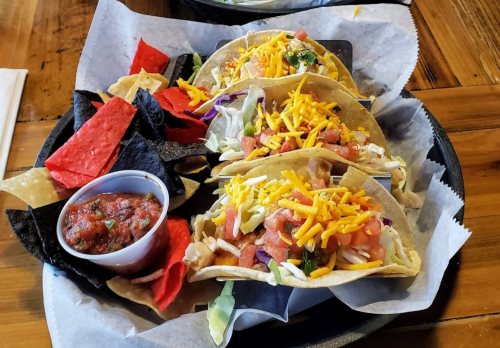 A plate of three tacos topped with cheese, lettuce, and tomatoes, served with salsa and colorful tortilla chips.