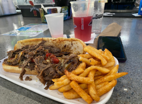 A plate with a beef sandwich topped with peppers and fries, alongside two drinks on a bar counter.