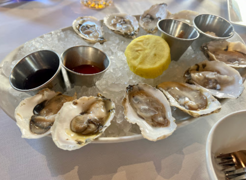 A platter of fresh oysters on ice, accompanied by two dipping sauces and a lemon wedge.