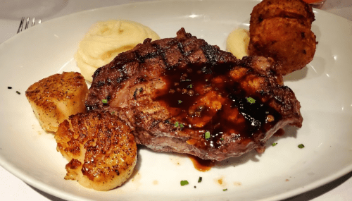 A plate featuring a grilled steak with sauce, mashed potatoes, and seared scallops, garnished with herbs.