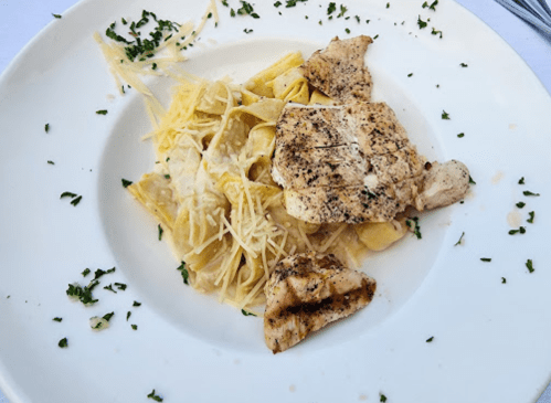 Grilled chicken served over creamy pasta with cheese, garnished with parsley on a white plate.