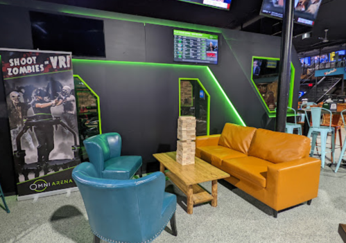 A cozy lounge area with a brown sofa, blue chairs, and a wooden table, featuring a zombie VR game poster on the wall.