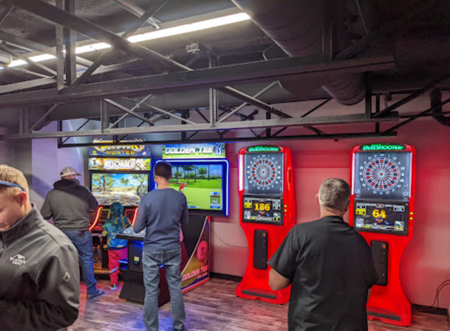 A game room with people playing arcade games and darts, featuring colorful machines and a modern atmosphere.