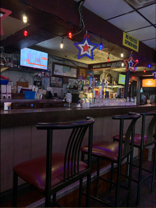 A dimly lit bar with empty stools, colorful lights, and a TV displaying a sports channel in the background.
