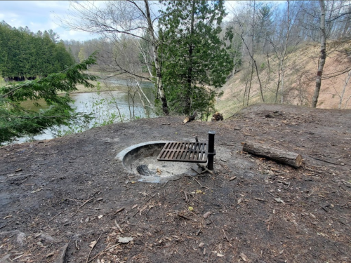 A fire pit with a metal grate sits on a dirt area near a river, surrounded by trees and a hillside.