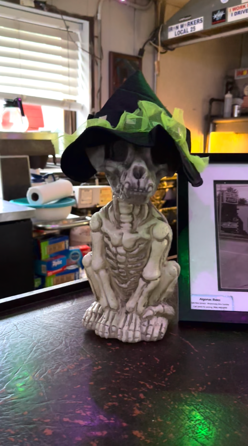 A skeleton figurine wearing a witch's hat sits on a bar counter, surrounded by colorful decorations.