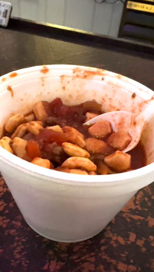 A white container filled with a chunky tomato-based dish, featuring pieces of chicken and vegetables, with a spoon inside.