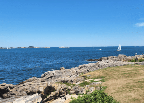 A serene coastal view featuring blue waters, rocky shoreline, and a sailboat under a clear blue sky.