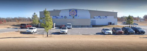 A large building with a colorful sign, surrounded by parked cars and trees, set in an open area.