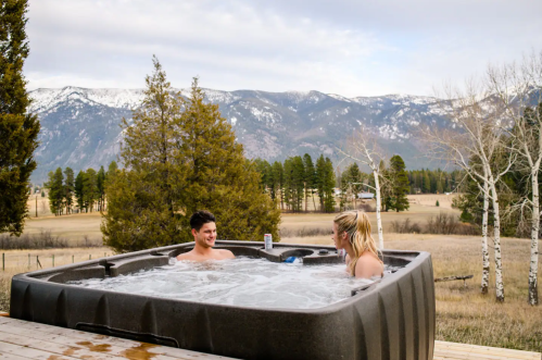 A man and woman relax in a hot tub with mountains and trees in the background, enjoying a scenic outdoor view.
