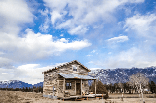 A rustic wooden house on a grassy field, surrounded by mountains and a blue sky with scattered clouds.