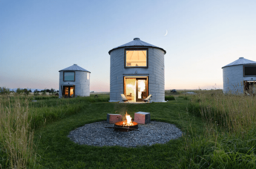 Two modern silos with large windows, a fire pit in front, and a serene sunset sky in the background.
