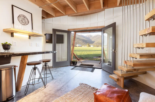 Cozy interior with open doors, wooden stairs, and a view of a grassy field and mountains in the background.