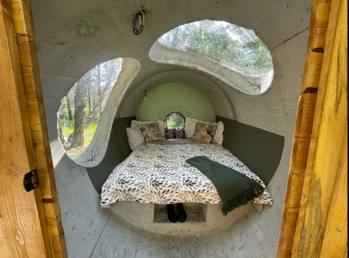 Cozy, circular interior of a unique pod-style room with a bed, pillows, and large windows surrounded by nature.