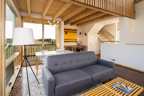 Modern living room with a gray sofa, wooden beams, and large windows overlooking a scenic view.