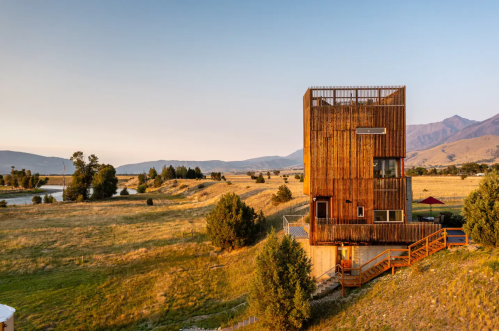 A modern wooden house on a hillside, surrounded by fields and mountains under a clear sky at sunset.