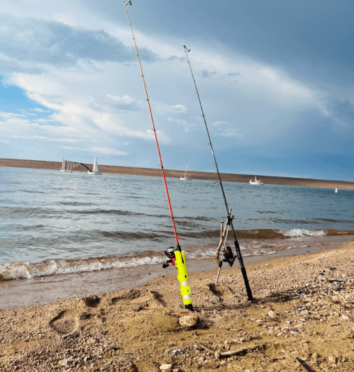 Two fishing rods stand in the sand by a lake, with sailboats visible in the background under a cloudy sky.