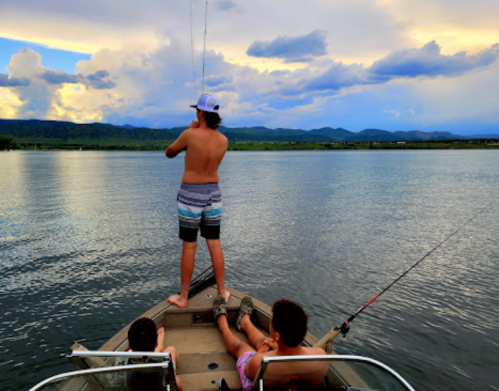 A shirtless person stands on a boat, fishing, while two others relax with fishing rods in a calm lake under a cloudy sky.