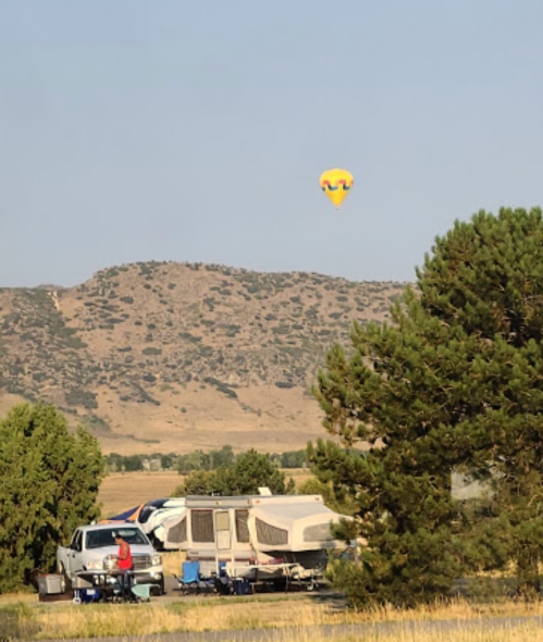 A hot air balloon floats above a campsite with a trailer and trees, set against a mountainous backdrop.