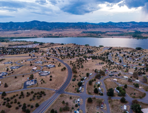 Aerial view of a lakeside area with winding roads, campsites, and mountains in the background under a cloudy sky.