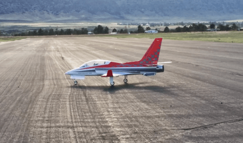 A small, red and silver model jet aircraft on a runway with a scenic landscape in the background.