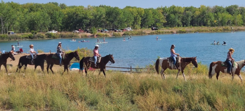 A group of people on horseback ride along a grassy shore by a lake, with kayakers visible in the background.