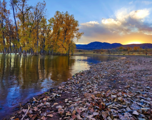A serene landscape featuring a calm river, autumn trees, and mountains under a colorful sunset sky.