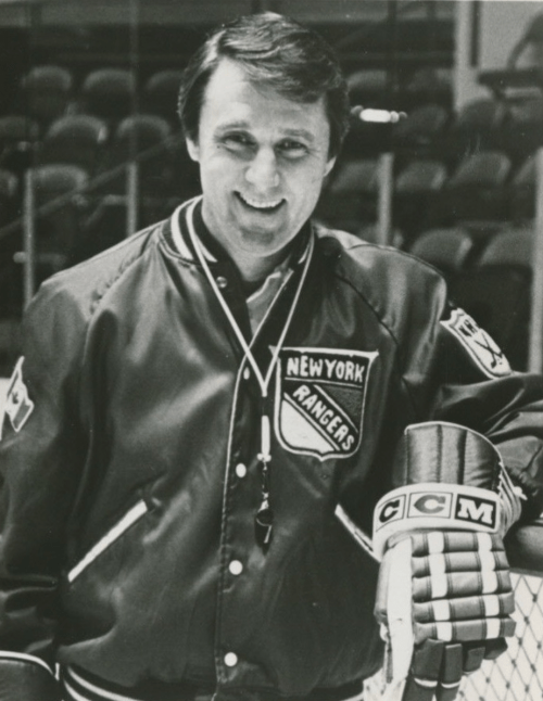 A smiling man in a New York Rangers jacket and gloves stands on an ice rink, holding a hockey stick.
