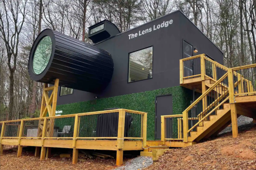 A modern black lodge with a unique cylindrical extension, surrounded by trees and featuring wooden decks.